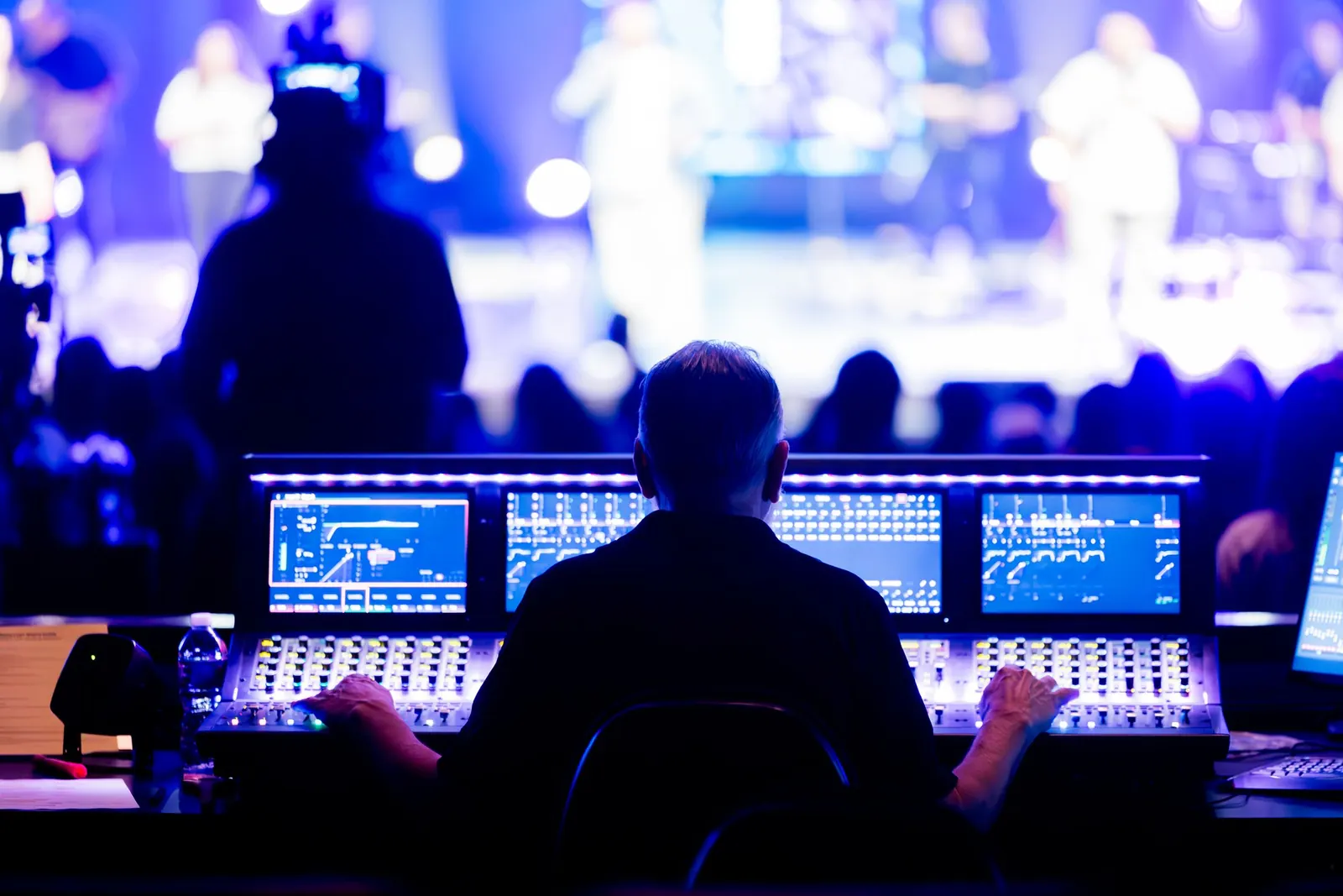 A tech volunteer at the sound desk in the booth, with screens and the lit worship stage beyond