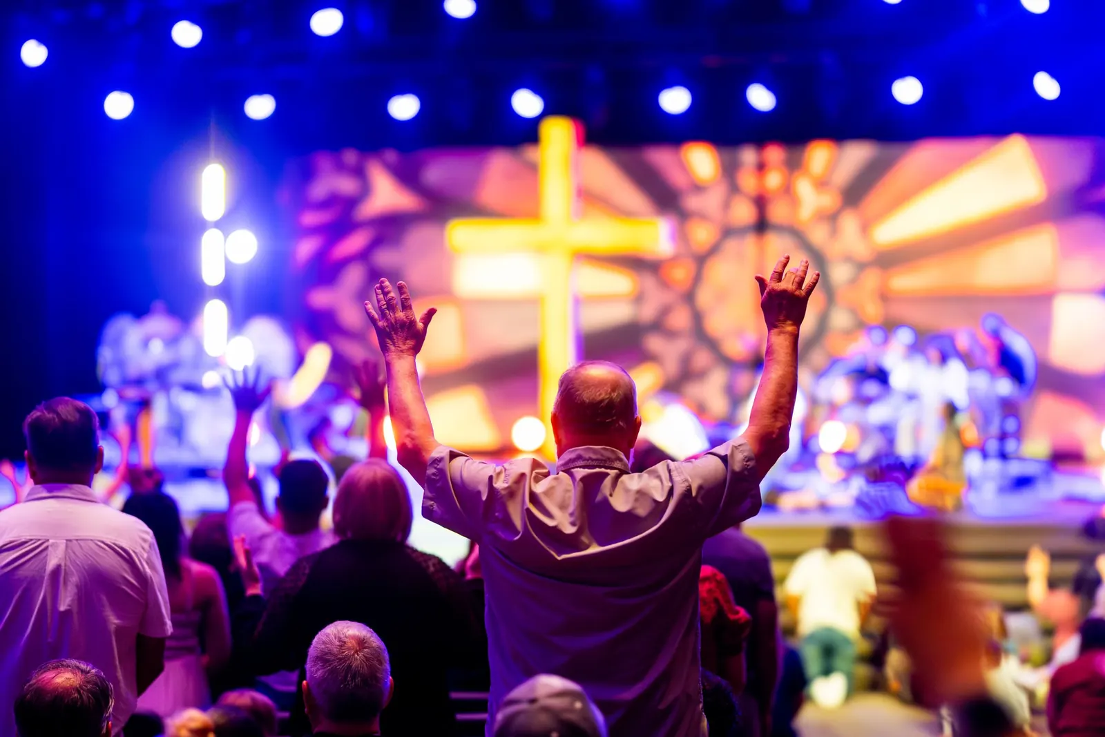 Worshippers with raised hands before a lit stage with a cross and large screens